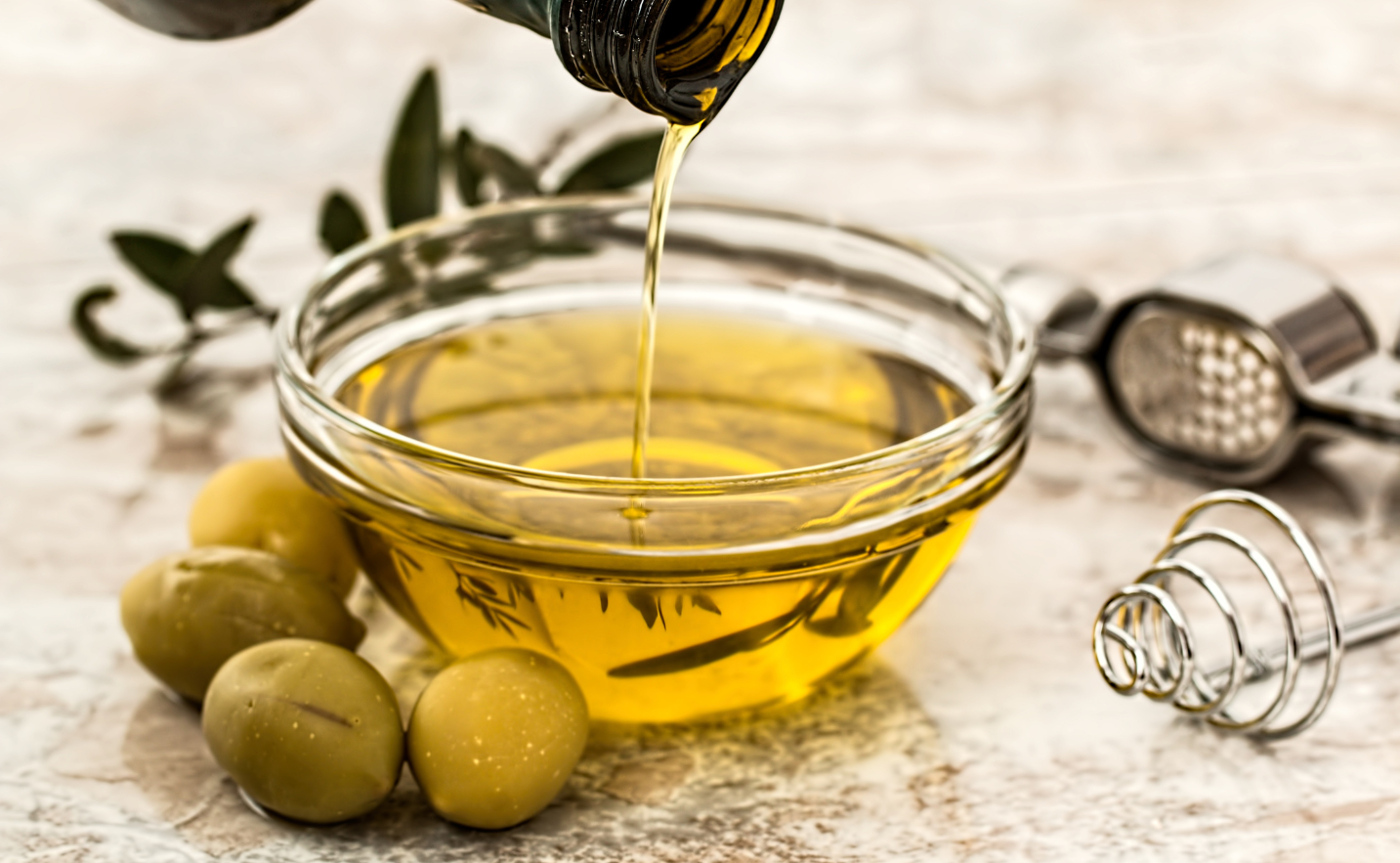 Olive oil being poured into a glass bowl with olives and a grater on a wooden surface.