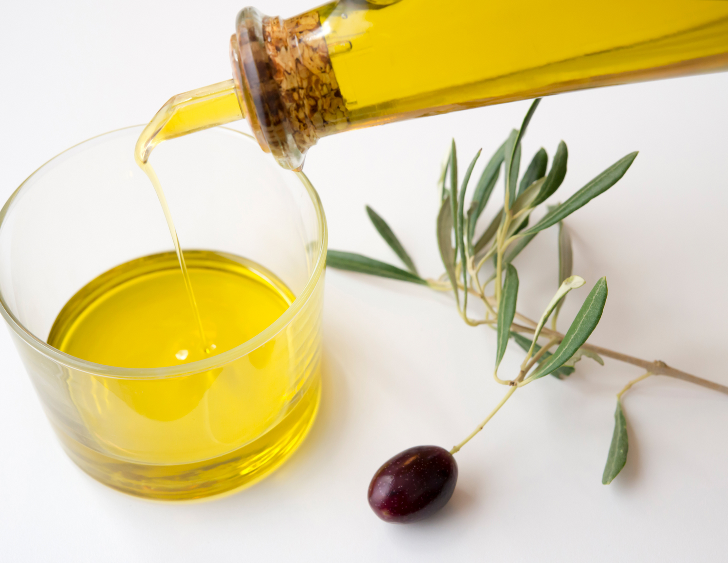 Olive oil being poured into a glass with an olive branch on a white background